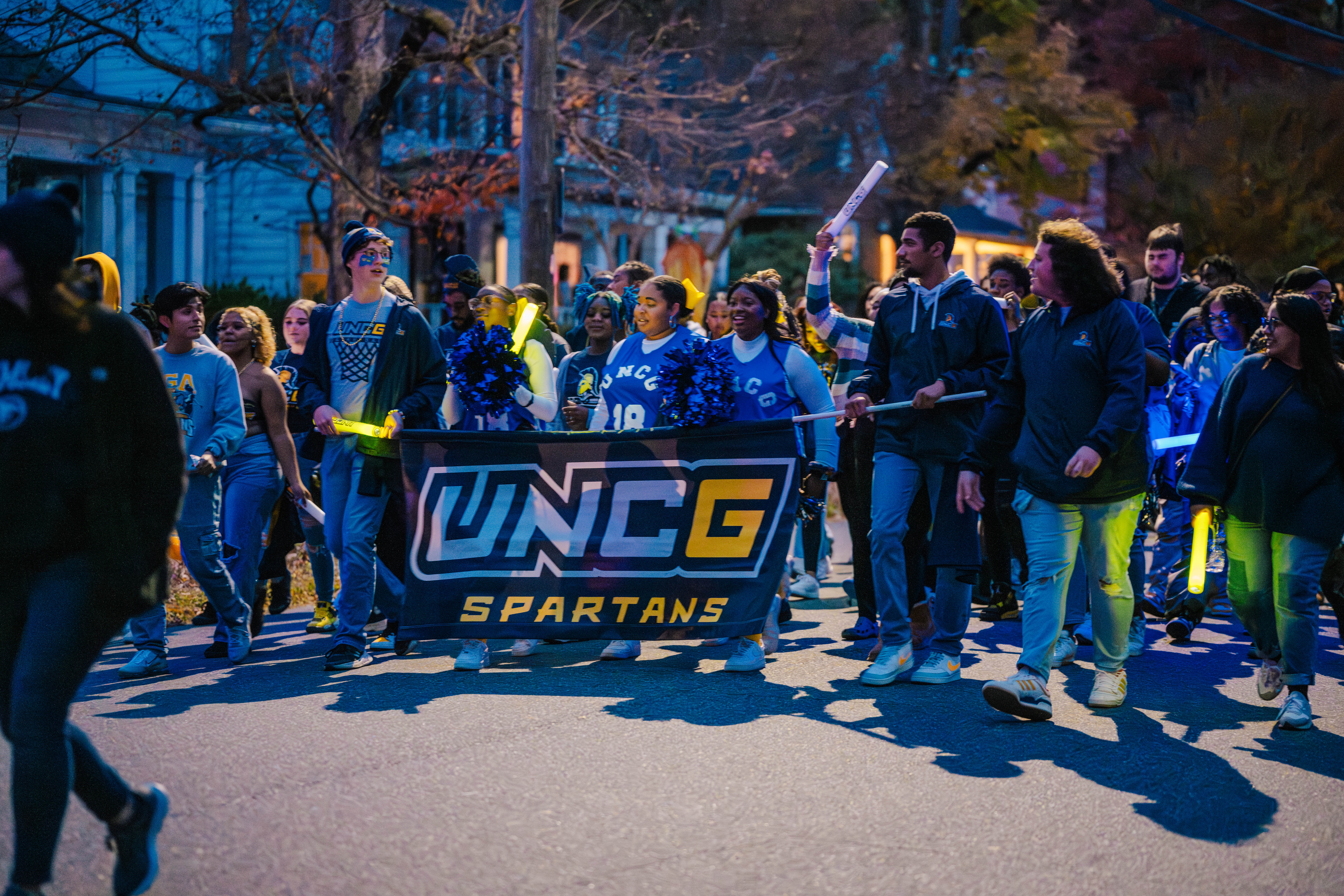Students march down a street at dusk holding a UNCG banner and cheering.