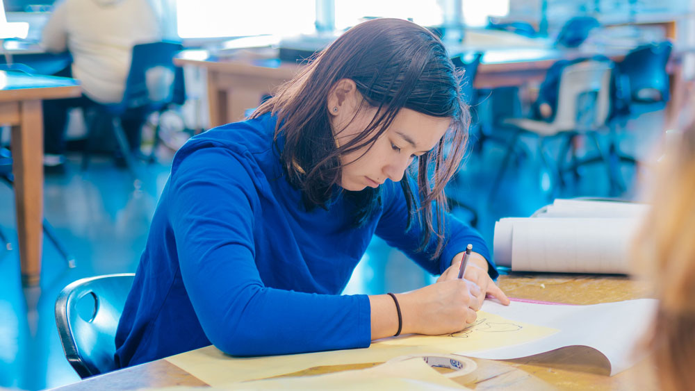 Students write on paper at a large classroom worktable.
