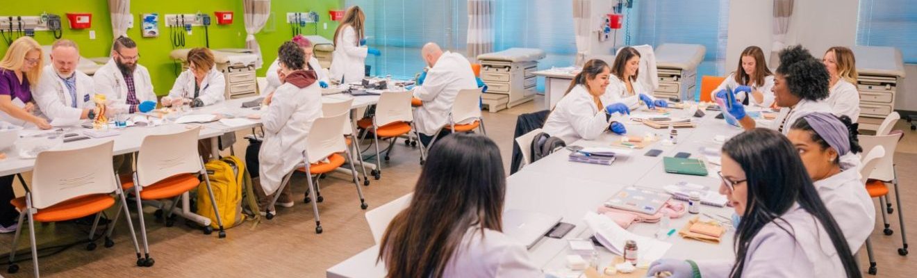 Nursing students in white lab coats practicing suturing at tables in a classroom together.