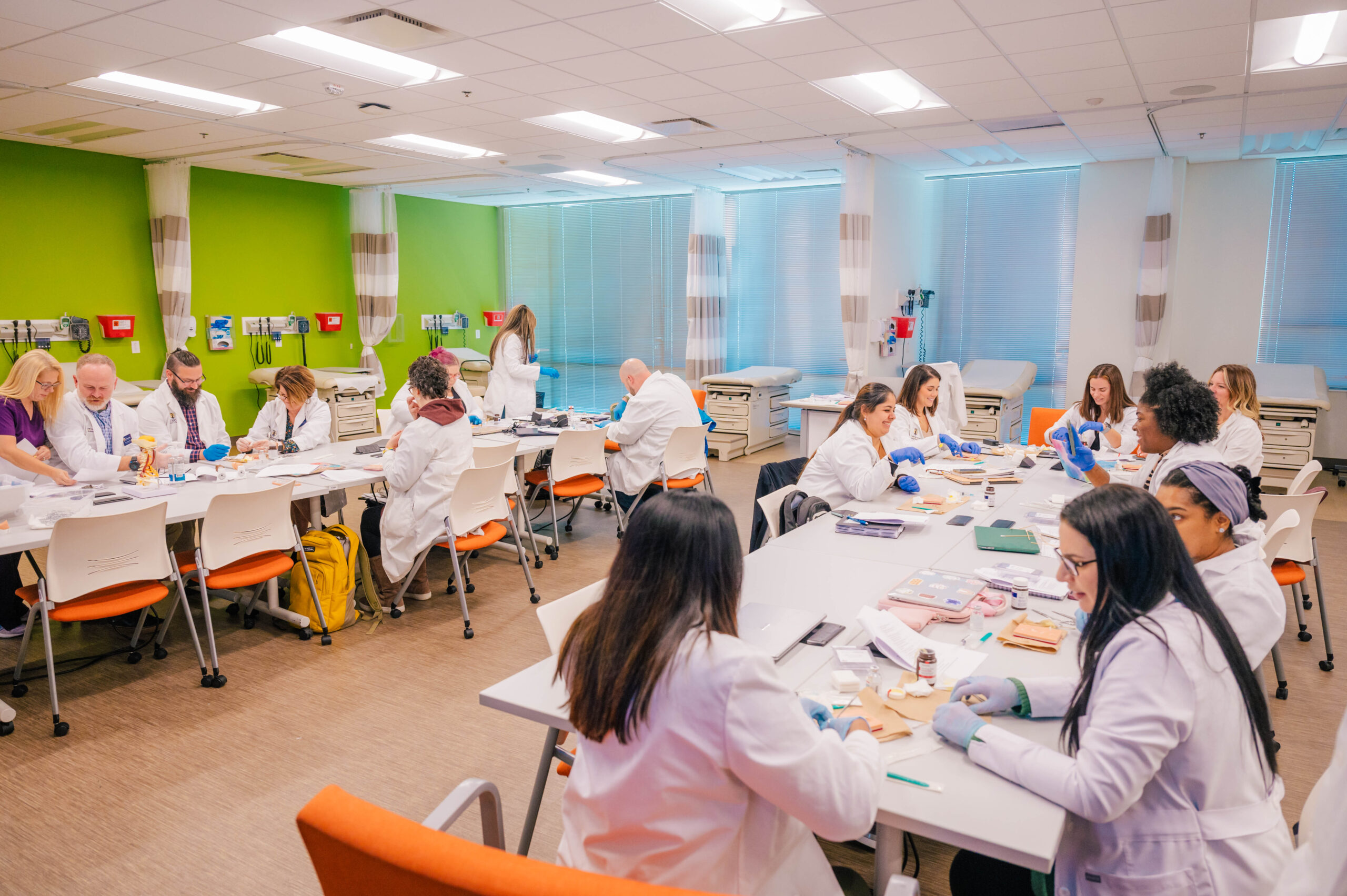 Nursing students in white lab coats practicing suturing at tables in a classroom together. 