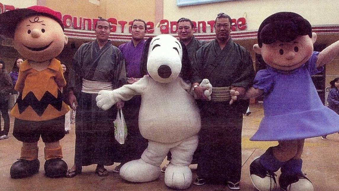 Sumo wrestlers pose with actors dressed as Charlie Brown, Snoopy, and Lucy.