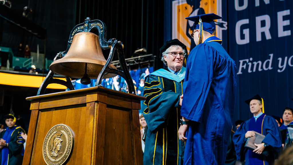 UNCG faculty and graduating student shake hands beside the bell at Commencement.