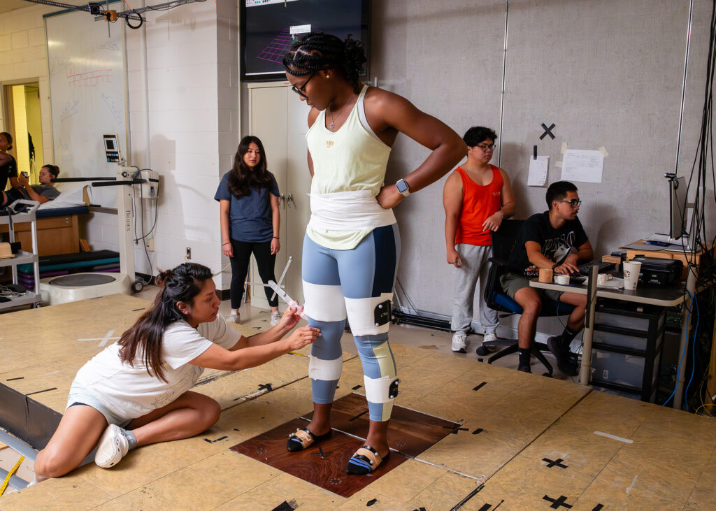 A woman in exercise clothes stands on a platform as a student attaches sensors to her legs. In the background others look at a computer monitor.