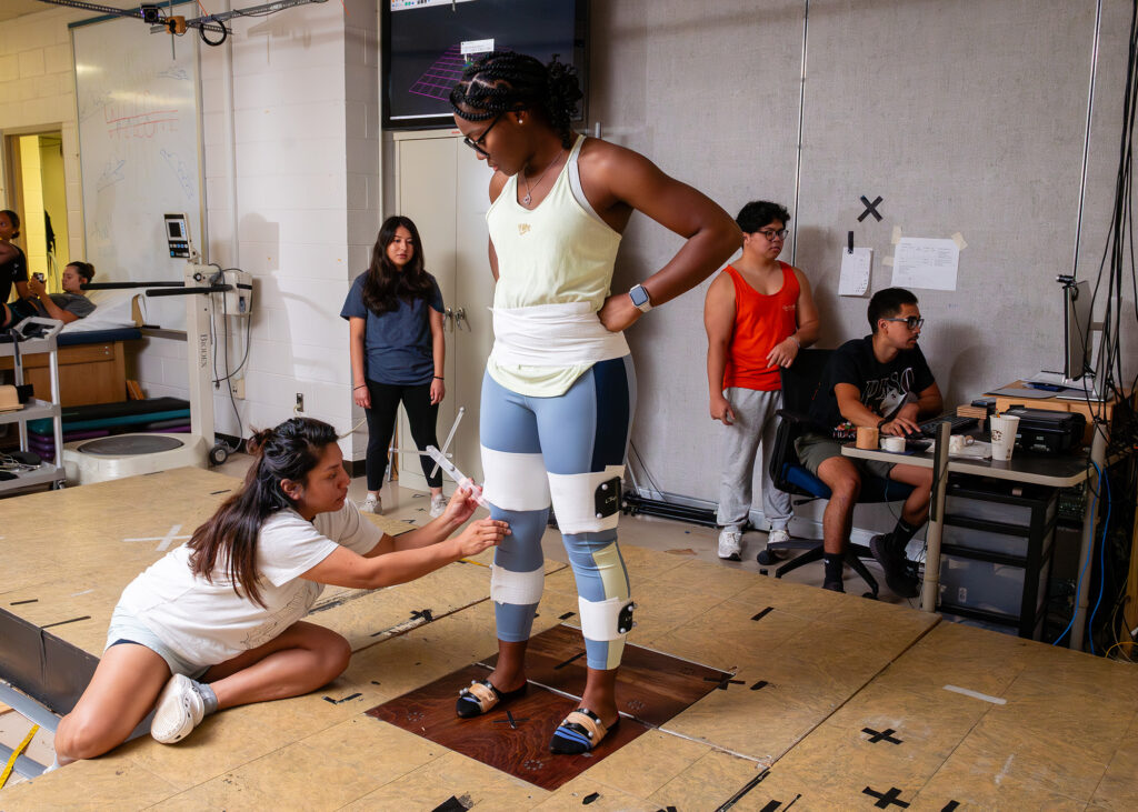 A woman in exercise clothes stands on a platform as a student attaches sensors to her legs. In the background others look at a computer monitor. 