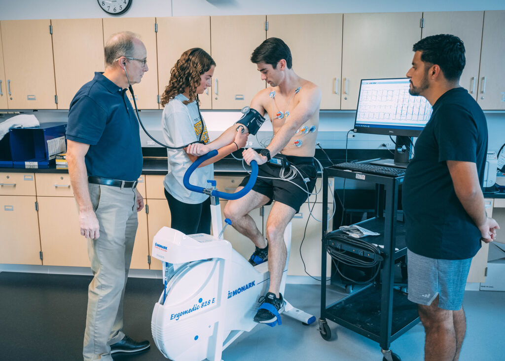 A man on a stationary bike gets his blood pressure taken as a teacher and two other students look on.
