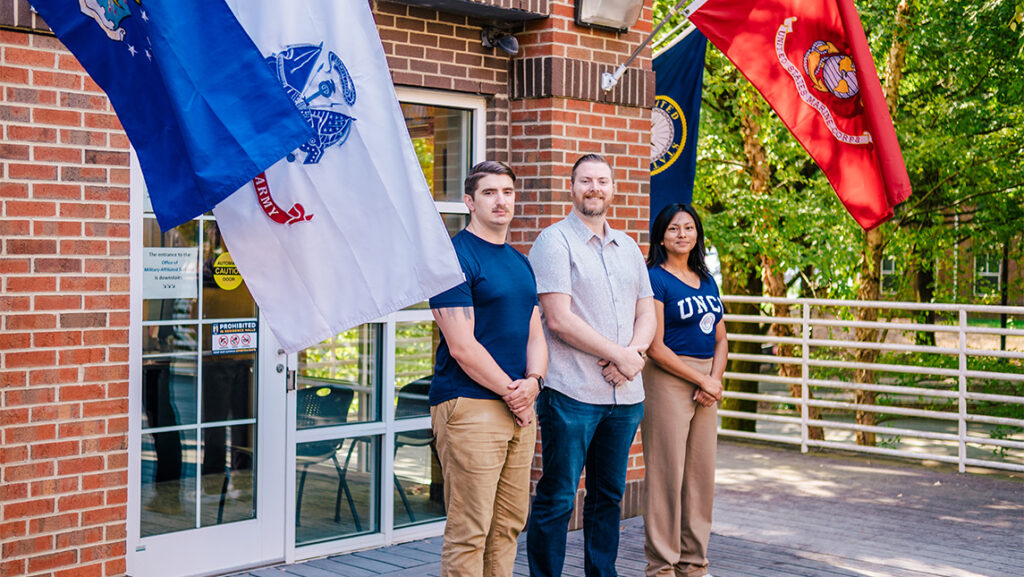 Three UNCG students stand beside the flags of their military branches.