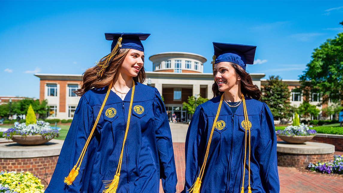 Two women in caps and gowns walk down the sidewalk in front of Moran Commons towards the camera.