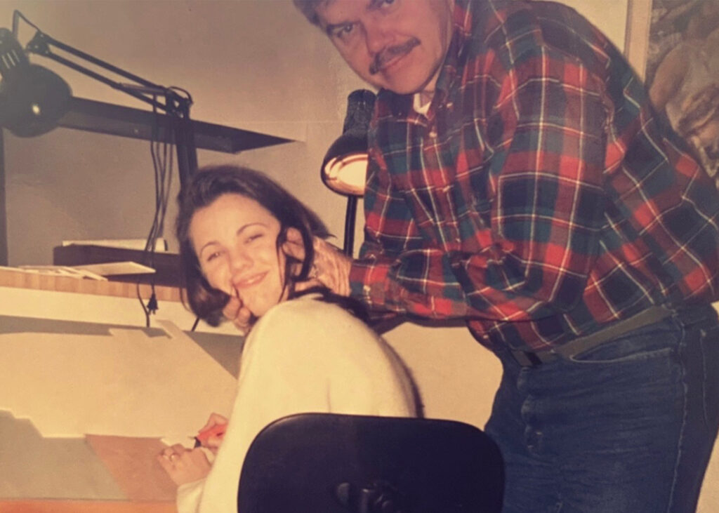 A young girl works at a drafting table with her dad behind her squeezing her face to smile for the camera.