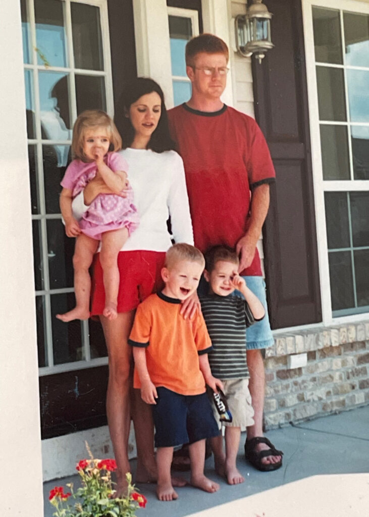 A young couple with 3 toddlers pose on the front porch of their house.
