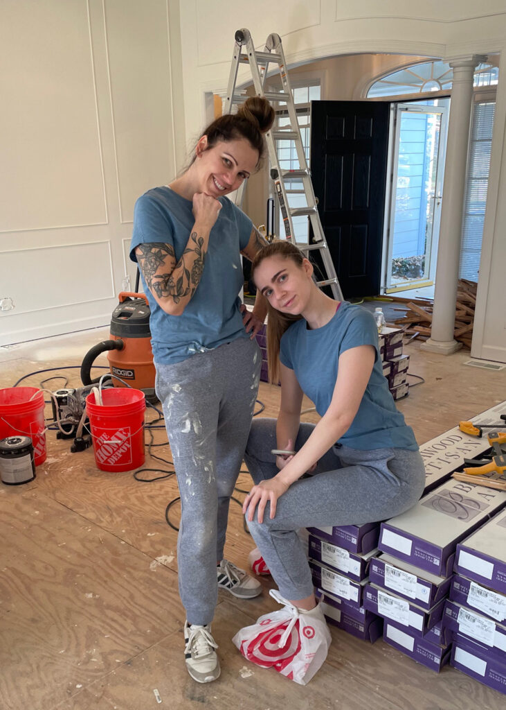 Two women stand in a room of a house they are renovating in paint stained clothes with a ladder and construction equipment around them.