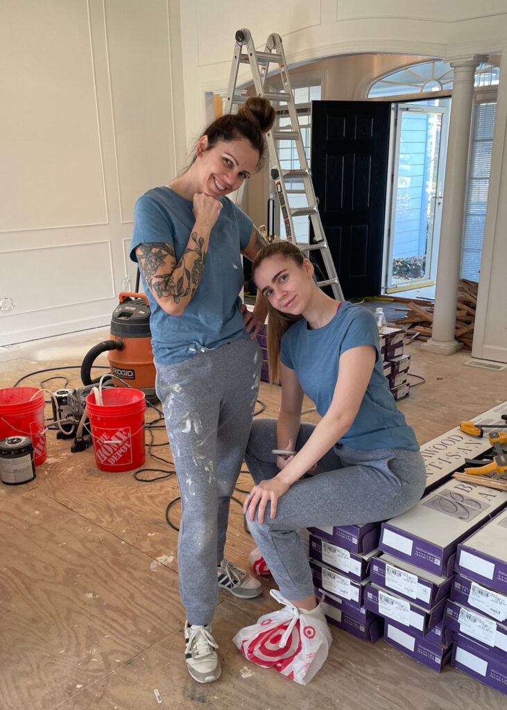 Two women stand in a room of a house they are renovating in paint stained clothes with a ladder and construction equipment around them. 