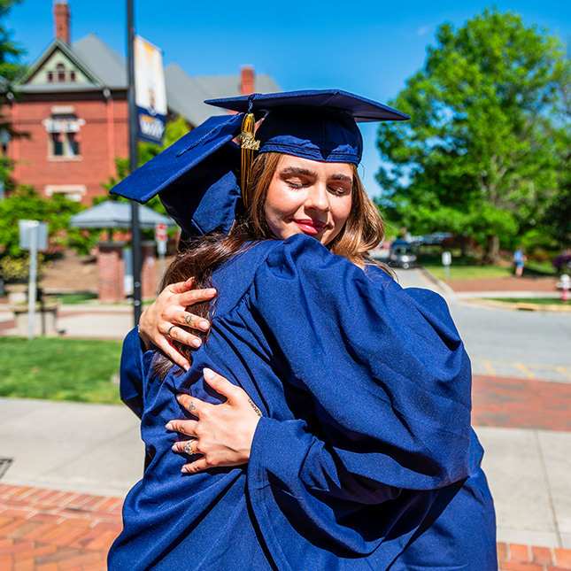 Two women dressed in caps and gowns hug in front of the Alumni House