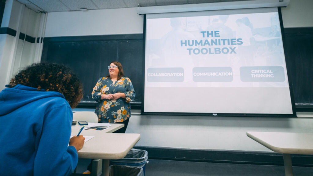 Professor stands in front of a class of students with a slide behind her with a "humanities toolbox" heading.