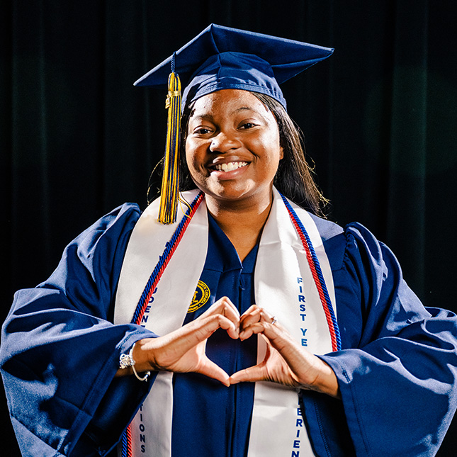 UNCG graduate poses in her cap and gown and makes a heart with her hands in front of her chest.