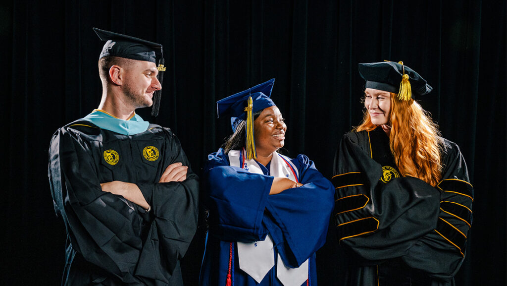 A master's, undergraduate and doctoral student pose in their caps and gowns with their arms crossed and smile at each other.