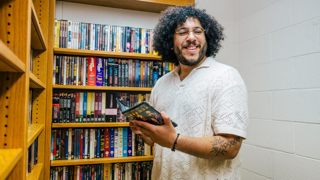 UNCG student stands among bookshelves with DVDs while holding a DVD of Iron Man