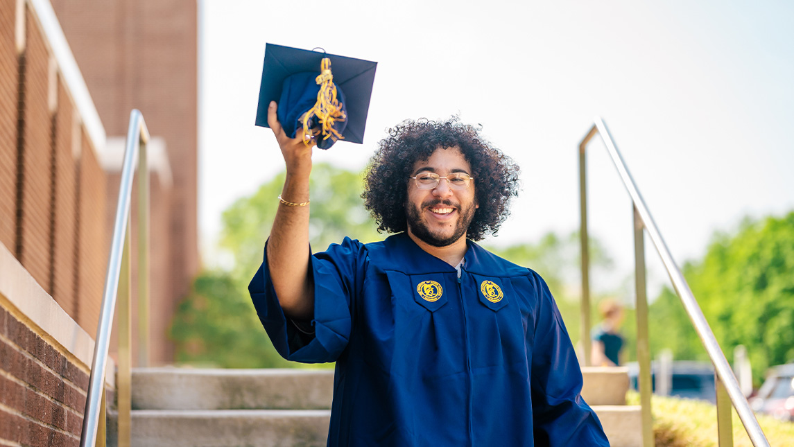 UNCG student in cap and gown smiling holding the cap