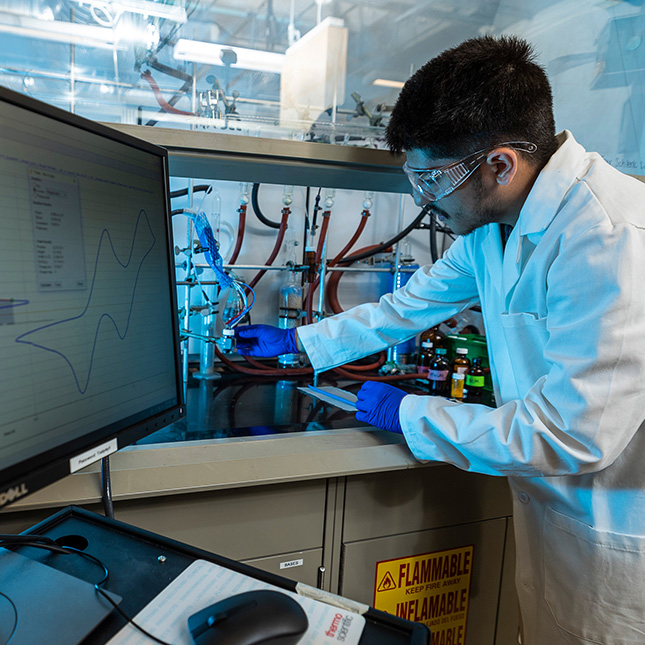 Student in lab coat works in a chemistry lab with a monitor screen showing graphs in the foreground.