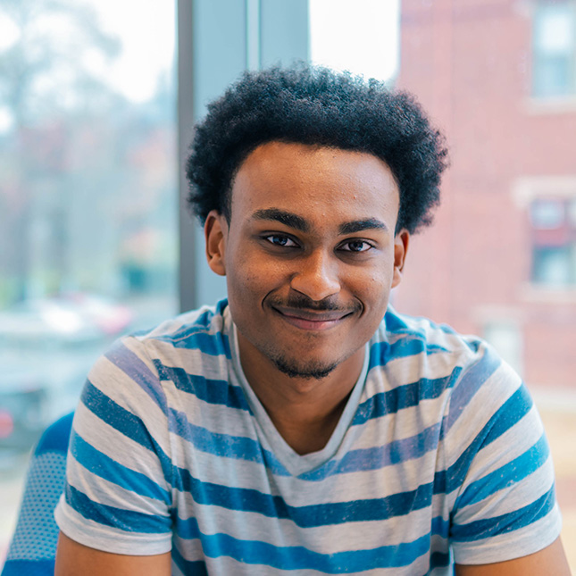 Student sits in a lounge in the Nursing Instruction Building with a confident smirk.