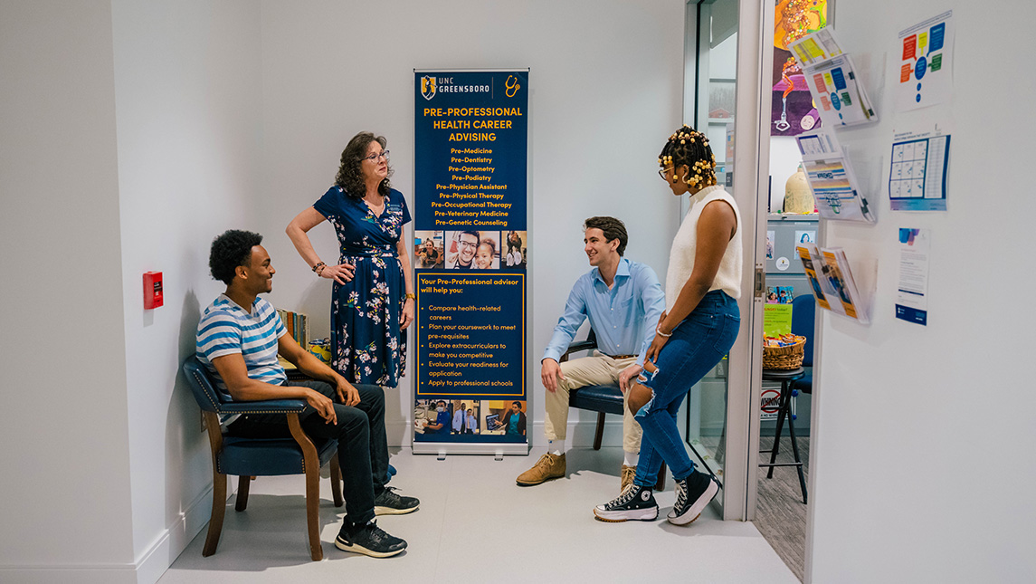 Three students gather in the hall with advisor, Robin Maxwell. Banner on wall behind them lists info about preprofessional programs.