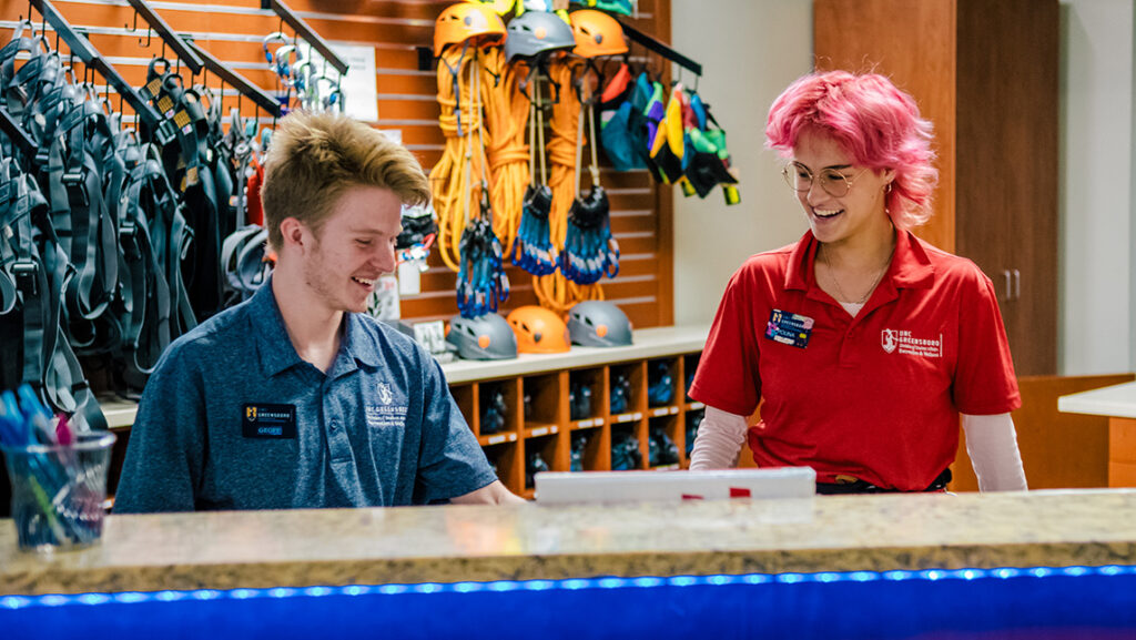 Two students work behind a desk at the Kaplan Center with harnesses and carabiners hanging on a wall behind them.
