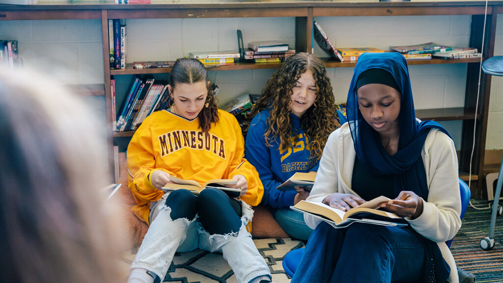 Three middle school students sit on a library floor reading books.