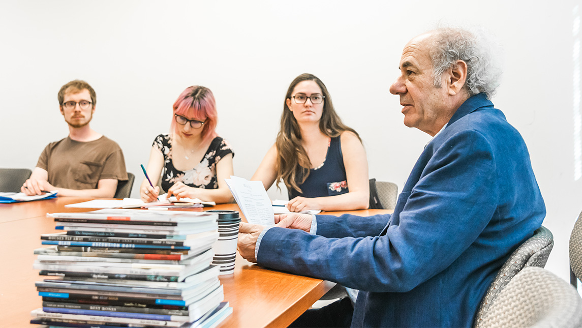 Author and UNCG professor Steve Dischell sits at a table with students and a stack of books.