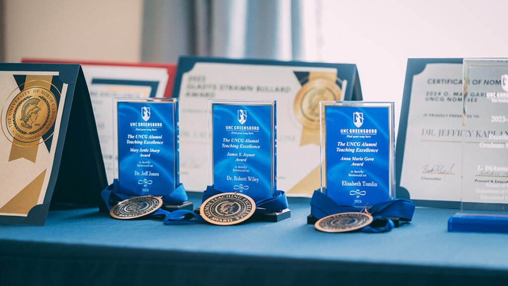 Awards, plaques, and medals for UNCG faculty sit on a table.