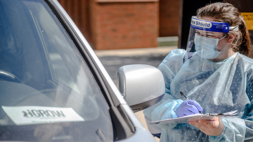 A woman in a mask and face shield with a clipboard stands beside a car's driver-side window at UNCG.
