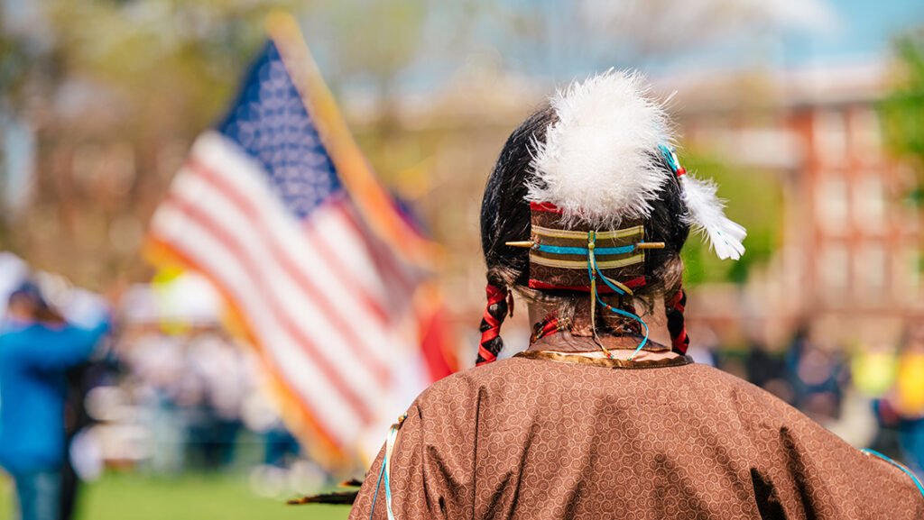 A Native American man in full regalia stands in front of the American flag on the UNCG campus.