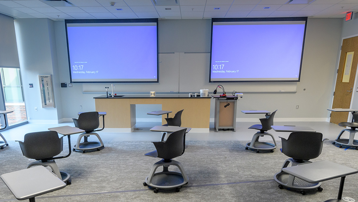 Chairs and desks clustered around the projector screens in a UNCG nursing classroom.