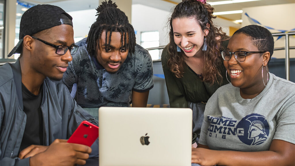 Students smile as they huddle around a laptop.