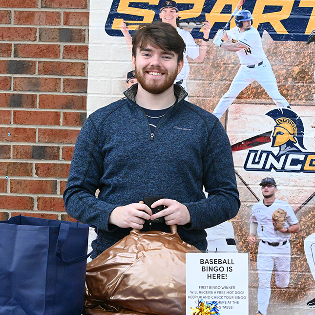 Man stands in front of a bring wall featuring UNCG baseball graphics and behind a table where he is handling marketing promotions.