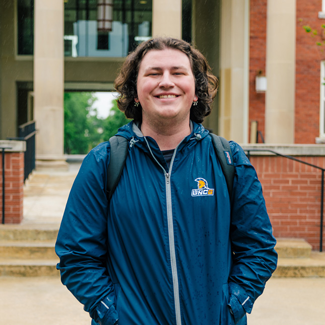Student stands in front of a residence hall in a UNCG branded jacket. and backpack.