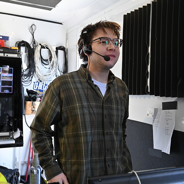 Student stands in a broadcast room with a headset on and wires hanging on the wall behind him.