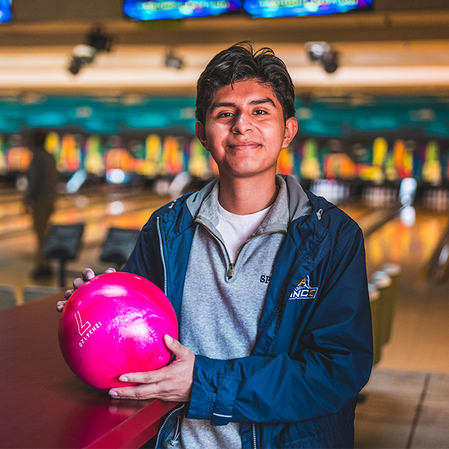 Student in a UNCG jacket stands in a bowling alley with a bowling ball.