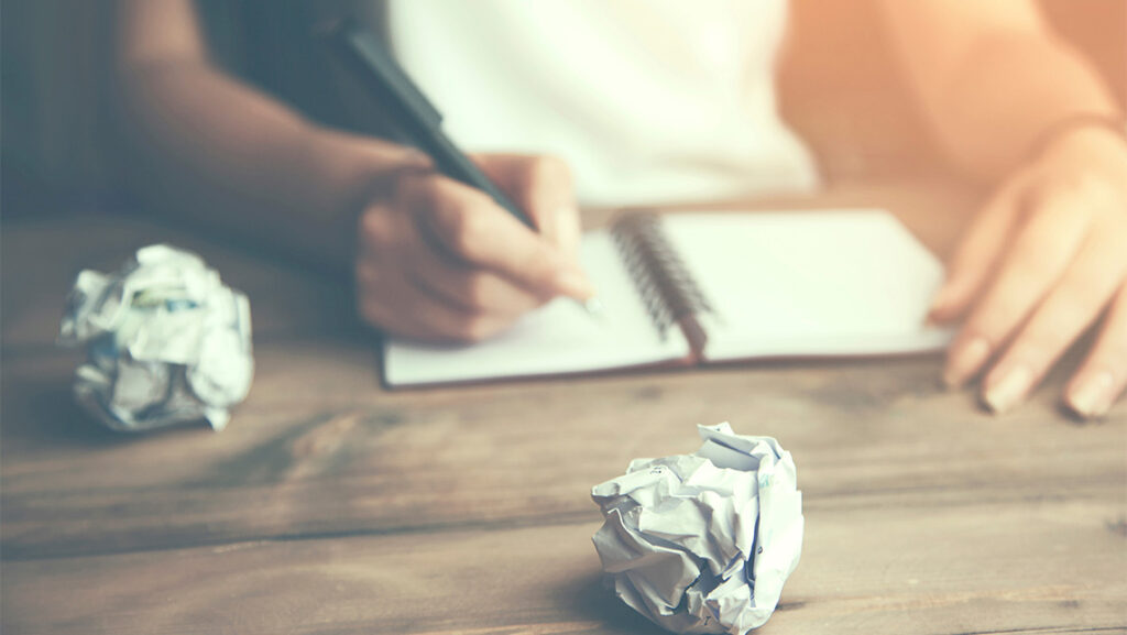 A person writing in a notebook surrounded by crumpled paper.