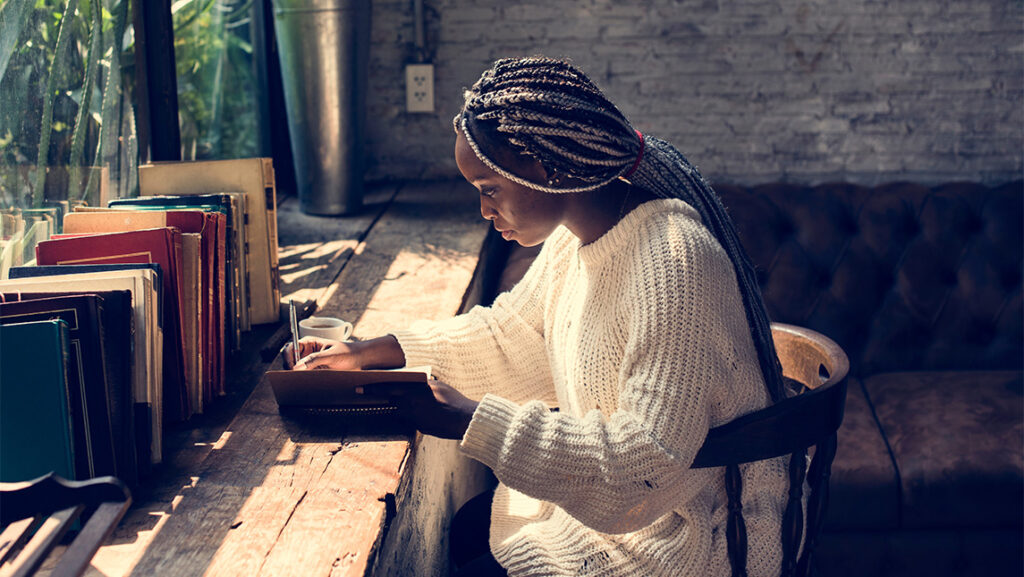 A woman sits at a table writing while surrounded by books.