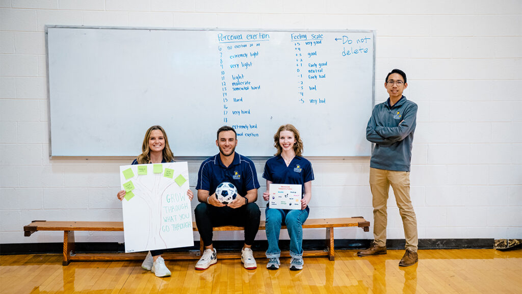Three students display their final projects while a professor stands next to them.