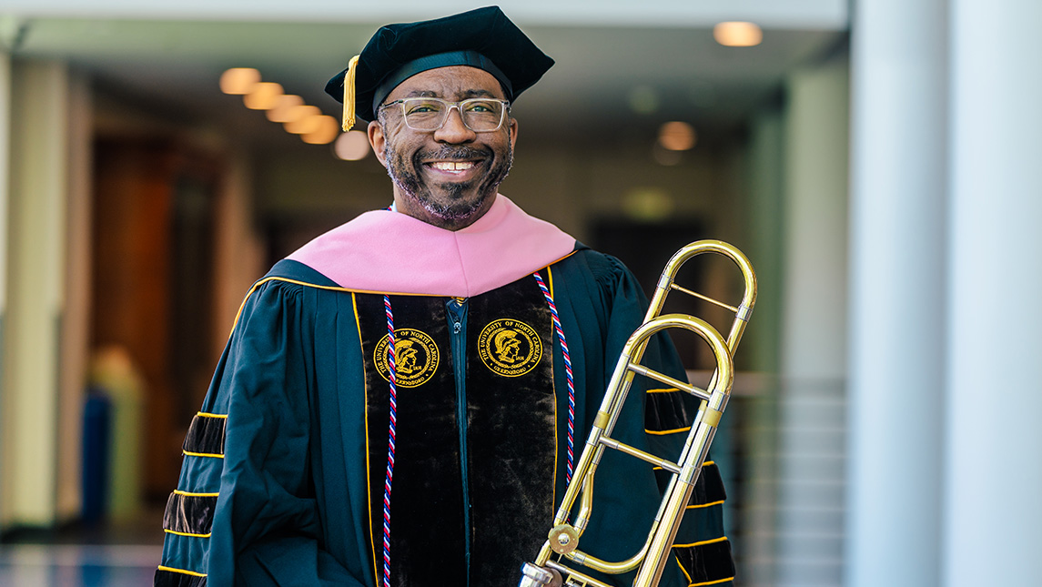 Dandrick Glenn stands with his trombone while wearing doctoral regalia