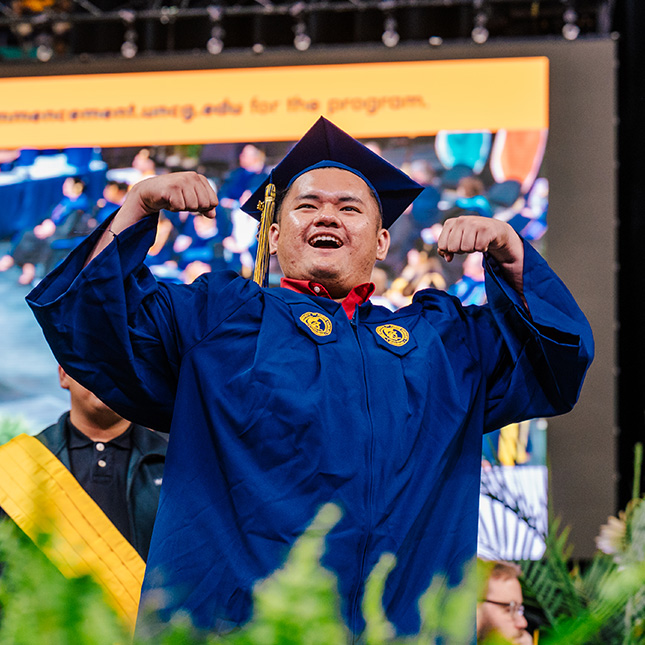 Graduate walks across stage in cap and gown with arms raised in a muscle flex pose.