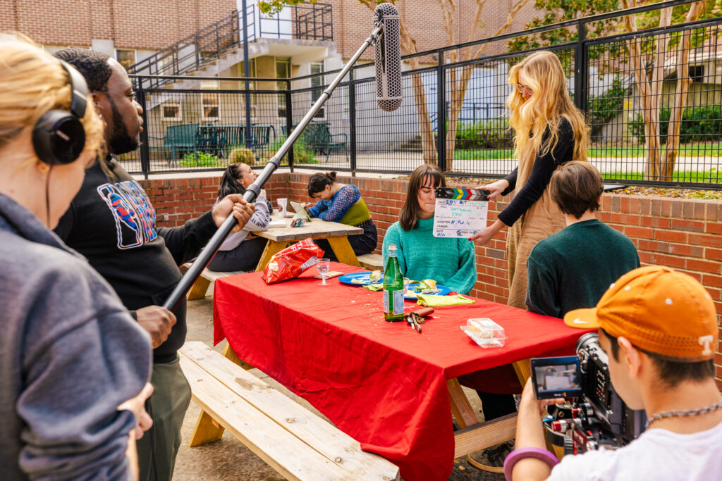 Students film a video at a picnic table on campus with a microphone and cameraman in the foreground. 