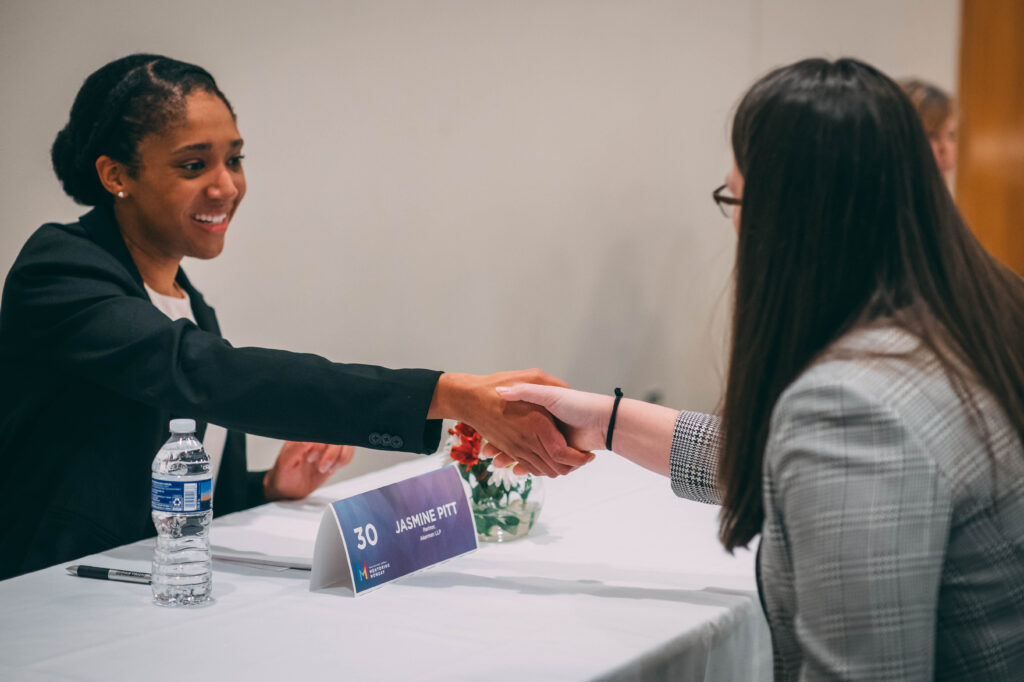 Women shake hands across a table at a mentoring event. 