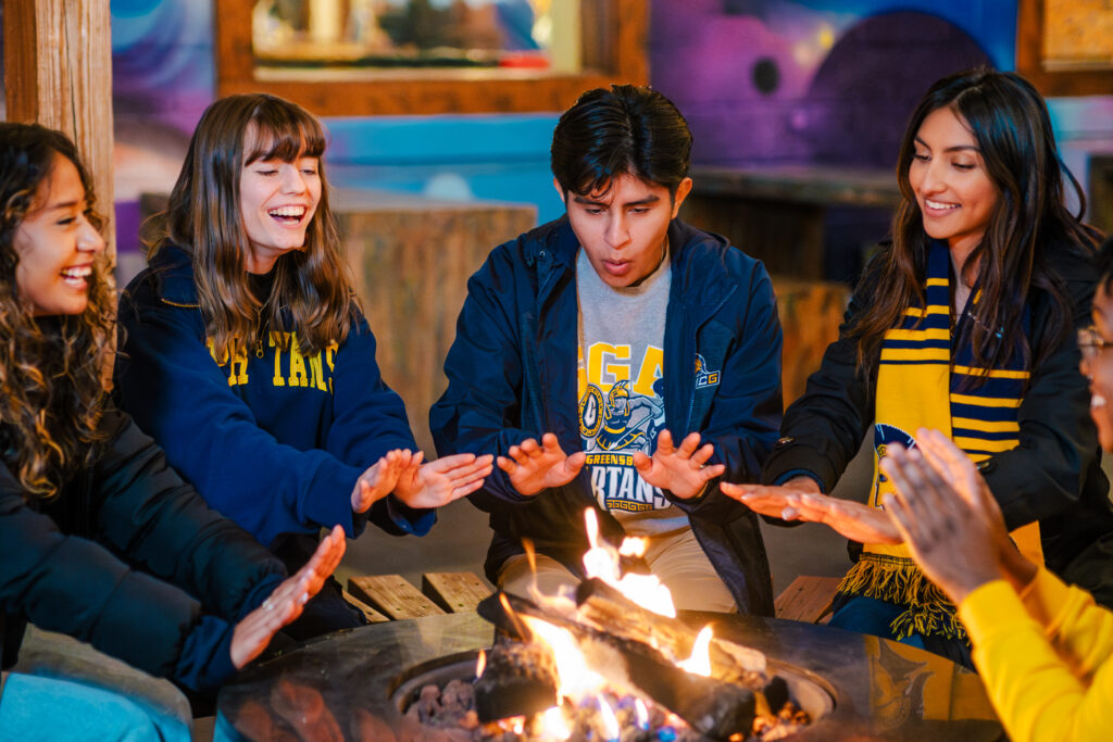 Students in UNCG sweatshirts gather around a firepit on a restaurant patio.
