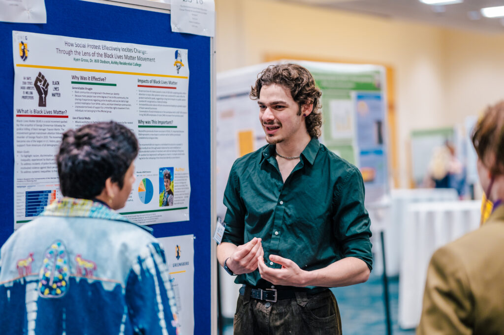 A student stands in front of a poster talking to another student with presentation boards in the background.