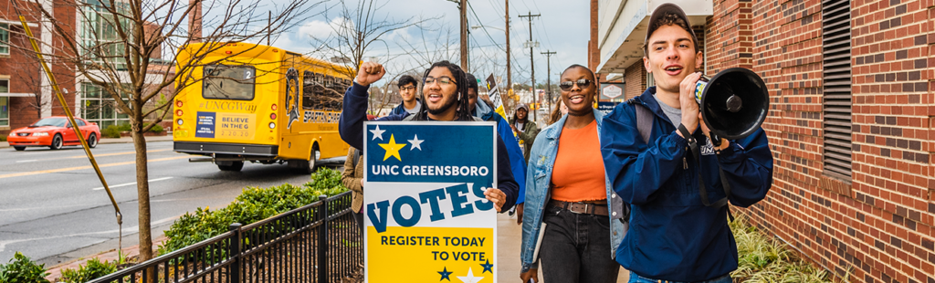 Students with megaphone and sign saying "UNC Greensboro VOTES" marching around campus
