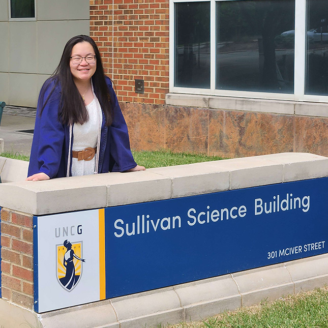 Student in graduation gown stands with hands on the Sullivan Science Building sign.