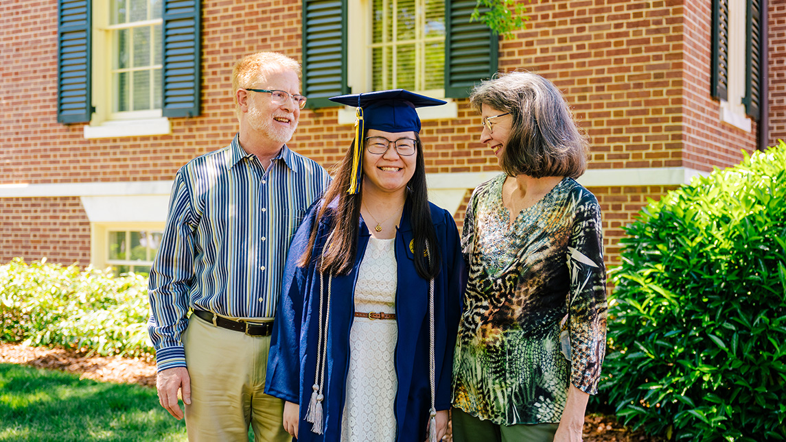 Student in cap and gown poses with parents on either side of her, looking at her proudly.