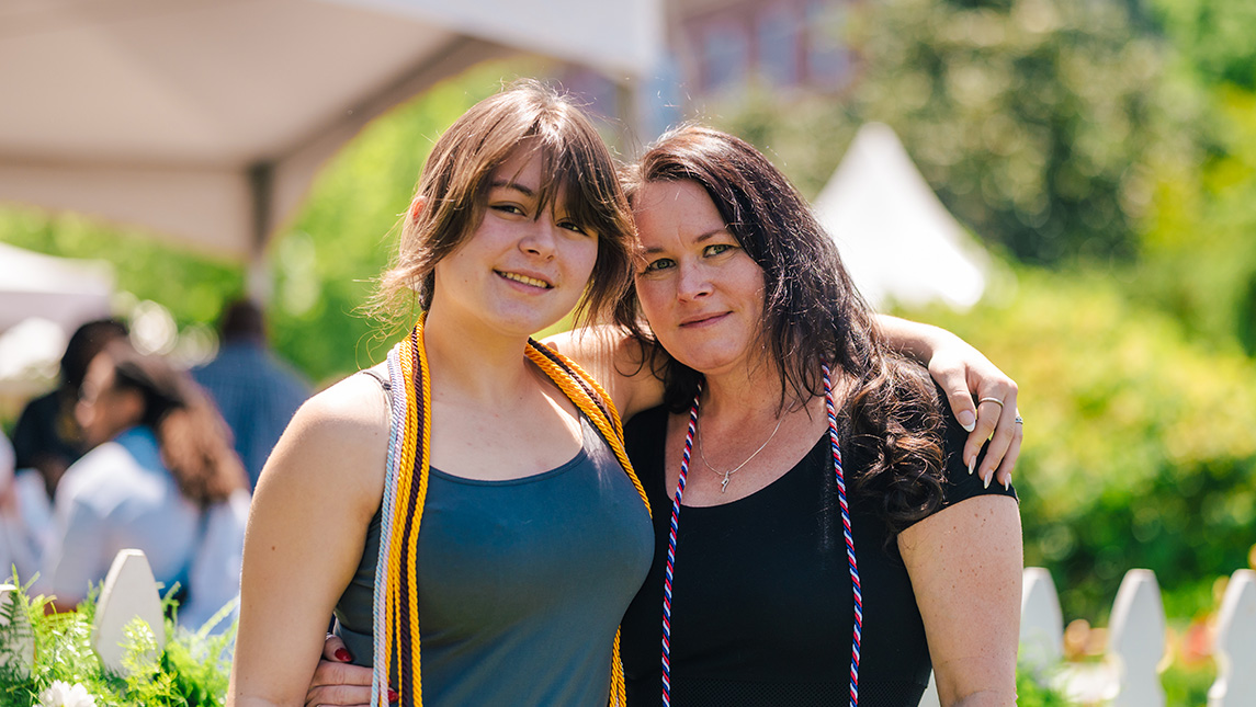 Mother and daughter pose on the UNCG campus with graduation honor cords.