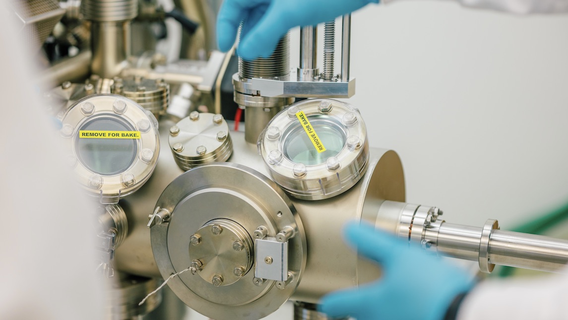 A pair of gloved hands work on a piece of machinery in the UNCG lab.